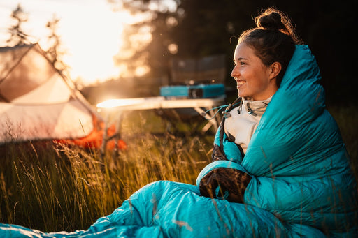 a woman outdoors by her tent wrapped in her sleeping bag